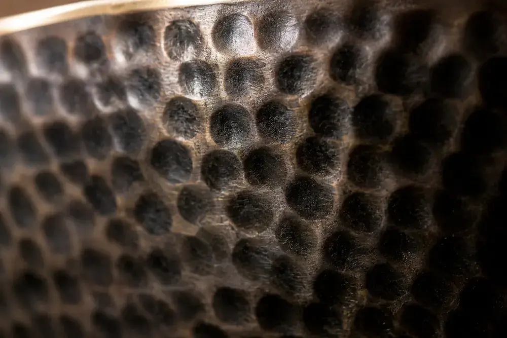 Extreme close-up of the dimpled hammer marks covering the darkened wall of a Nadam singing bowl, with the bright gold rim edge along the top